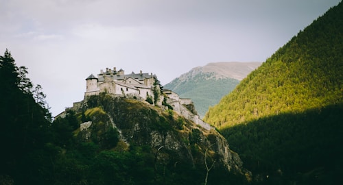 A fortress or castle sits atop a rocky hill surrounded by dense green forests and towering mountain landscapes. The architecture is classic and sturdy, with numerous towers and a light-colored facade that contrasts with the lush vegetation. Light filters through the trees, casting shadows across the scene, hinting at a tranquil yet imposing atmosphere.