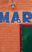 An orange brick wall with the letters 'MAR' painted in blue. A green door is partially visible on the right side, framed in red. There is also a light fixture above the letters.