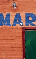 An orange brick wall with the letters 'MAR' painted in blue. A green door is partially visible on the right side, framed in red. There is also a light fixture above the letters.