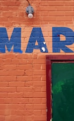 An orange brick wall with the letters 'MAR' painted in blue. A green door is partially visible on the right side, framed in red. There is also a light fixture above the letters.