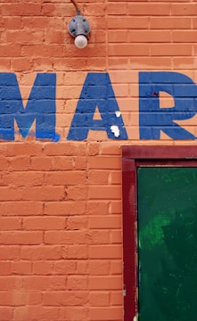 An orange brick wall with the letters 'MAR' painted in blue. A green door is partially visible on the right side, framed in red. There is also a light fixture above the letters.