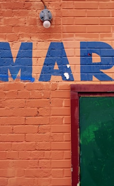 An orange brick wall with the letters 'MAR' painted in blue. A green door is partially visible on the right side, framed in red. There is also a light fixture above the letters.