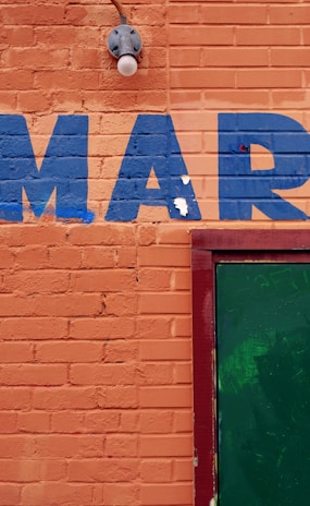 An orange brick wall with the letters 'MAR' painted in blue. A green door is partially visible on the right side, framed in red. There is also a light fixture above the letters.
