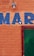 An orange brick wall with the letters 'MAR' painted in blue. A green door is partially visible on the right side, framed in red. There is also a light fixture above the letters.