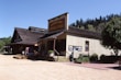 A vintage general store with a wooden facade, adorned with signs and surrounded by trees and shrubs. The building has a rustic appearance with a steeply pitched roof and a large, prominent sign. Nearby, bales of hay are arranged in a stack, and a small structure resembles a booth or playhouse.