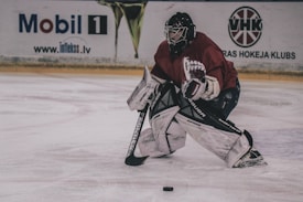 A hockey goalie is positioned on an ice rink, wearing full protective gear including a helmet, leg pads, gloves, and a blocker. The player is crouched in a defensive stance, focused on the hockey puck on the ice near their skates. Advertising boards are visible in the background.