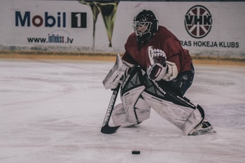 A hockey goalie is positioned on an ice rink, wearing full protective gear including a helmet, leg pads, gloves, and a blocker. The player is crouched in a defensive stance, focused on the hockey puck on the ice near their skates. Advertising boards are visible in the background.