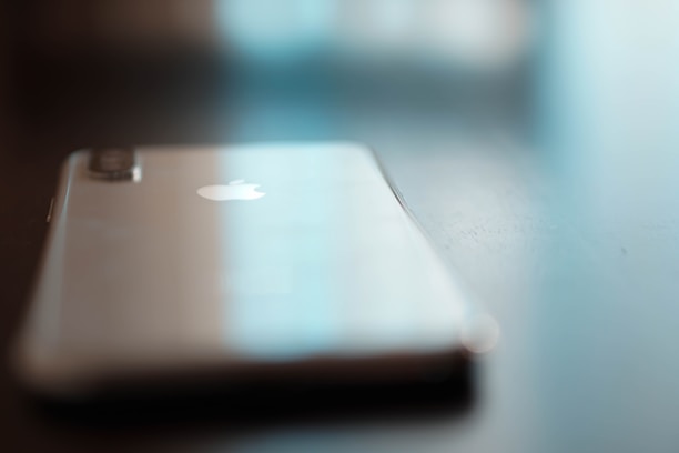 A smartphone lies on a reflective surface, slightly out of focus. The metallic back and Apple logo are visible, with soft lighting creating a serene and modern feel.