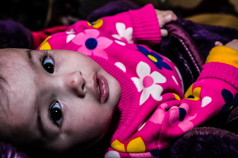 A baby wearing a bright pink sweater with colorful floral patterns looks up with a calm expression. The sweater features large flowers in white, pink, and yellow colors. The baby’s eyes are wide open, and they are lying on what appears to be a dark, soft blanket.