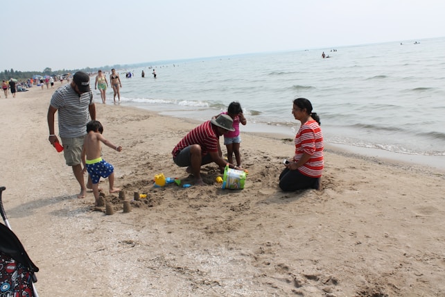A group of people is engaged in building sandcastles on a sandy beach. Two adults and two children are focused on their play, with sand buckets and shovels scattered around. The beach is populated with other people, some walking and some enjoying the water. The ocean is calm, stretching into the distance under a clear sky.