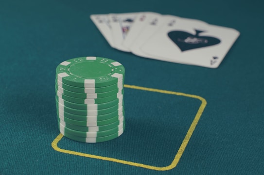 A stack of green poker chips is positioned on a green felt table within a yellow rectangular outline. In the background, there is a fanned set of playing cards, including an ace of spades visible.