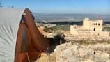 A campsite on rocky terrain features a large tent with an orange and gray exterior. In the middle distance, a historic-looking stone fortress with a red flag stands atop a hill, overlooking a sprawling city or townscape with mountains in the background.