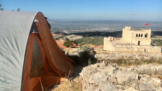 A campsite on rocky terrain features a large tent with an orange and gray exterior. In the middle distance, a historic-looking stone fortress with a red flag stands atop a hill, overlooking a sprawling city or townscape with mountains in the background.