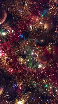 A close-up view of a Christmas tree decorated with colorful lights, vibrant garlands, and shiny baubles. The ornaments reflect the surrounding lights, creating a festive and warm ambiance.