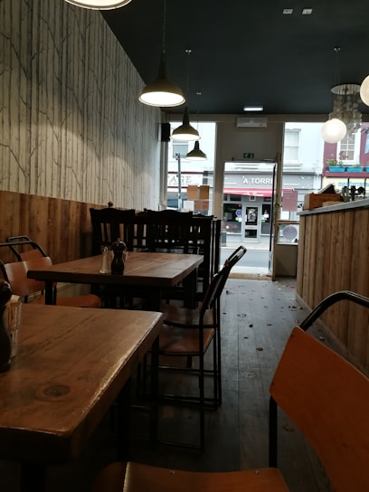The interior of a cozy café features wooden tables and chairs in a narrow space with dim lighting. The walls have a rustic wood paneling on the lower half and a tree-patterned wallpaper above. Hanging pendant lights provide illumination, and the view towards the glass door reveals part of a street outside.
