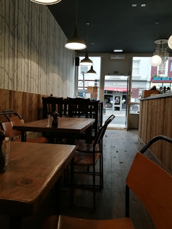 The interior of a cozy café features wooden tables and chairs in a narrow space with dim lighting. The walls have a rustic wood paneling on the lower half and a tree-patterned wallpaper above. Hanging pendant lights provide illumination, and the view towards the glass door reveals part of a street outside.