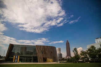 A modern building with reflective glass and geometric design sits in the foreground, surrounded by a lush green lawn. In the background, a tall, pointed skyscraper and other city buildings rise against a sky filled with scattered clouds and a deep blue hue.