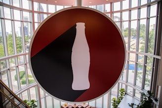 A large circular sign featuring a silhouette of a classic soda bottle is prominently displayed in front of an expansive, curved glass wall. The background of the sign is divided into red and black sections, and the silhouette is white. The surrounding area outside the glass wall includes greenery and buildings, suggesting an urban setting.