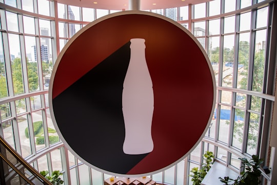 A large circular sign featuring a silhouette of a classic soda bottle is prominently displayed in front of an expansive, curved glass wall. The background of the sign is divided into red and black sections, and the silhouette is white. The surrounding area outside the glass wall includes greenery and buildings, suggesting an urban setting.