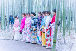 A group of women wearing colorful kimonos and shawls stand together in a bamboo forest. The kimonos feature vibrant patterns like flowers and stripes, and the scene is set on a path surrounded by tall bamboo stalks.