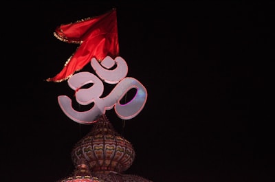 A large, illuminated Om symbol is positioned atop an ornate, dome-like structure, with intricate patterns and vibrant colors. A red flag with gold embellishments is prominently displayed, fluttering above the Om symbol against a dark background.