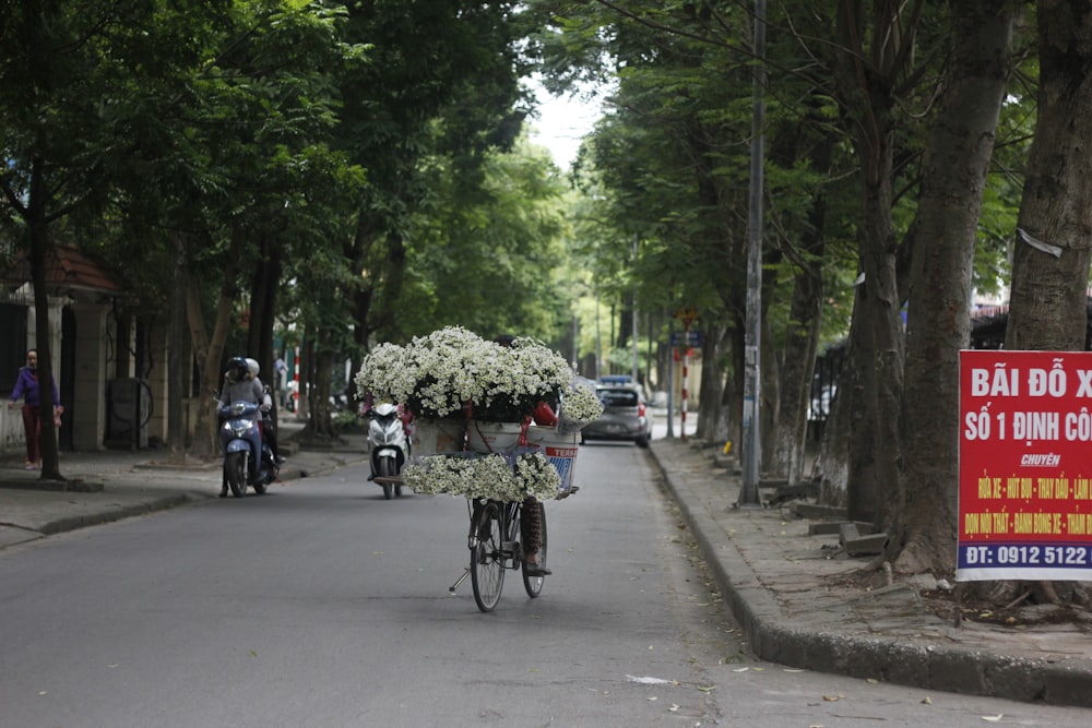 白い花を籠に入れて自転車に乗った人が木々の間の道を走っている