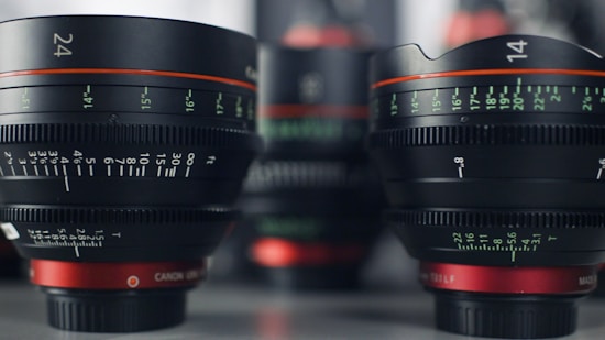 Several camera lenses, primarily focusing on two large lenses with visible focal length markings like '24' and '14'. The lenses are predominantly black with red accents and detailed measurement indicators in green and white.