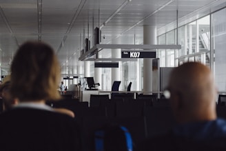 An airport gate area featuring empty seats, clear signage indicating Gate K07, and a modern architectural design with large windows. The foreground includes two blurred individuals, suggesting the image was taken candidly from behind them.
