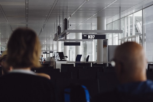 An airport gate area featuring empty seats, clear signage indicating Gate K07, and a modern architectural design with large windows. The foreground includes two blurred individuals, suggesting the image was taken candidly from behind them.