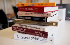 A stack of business and entrepreneurship books on a table. The titles are oriented towards strategies, innovation, and startup guidance, including 'Zero to One', 'Ego is the Enemy', and 'The Startup Owner's Manual'. The setting appears to be in a modern office or study environment.