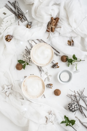 Two frothy cups of coffee are placed on a white textured table surrounded by rustic elements like cinnamon sticks, pine cones, and twigs. Decorative white fabric, small candles, and white star ornaments add to the aesthetic. Hints of green leaves and white berries are also present.