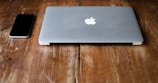 A closed, silver laptop with an apple logo next to a black smartphone on a rustic wooden table.