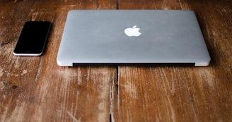 A closed, silver laptop with an apple logo next to a black smartphone on a rustic wooden table.