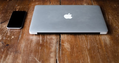 A closed, silver laptop with an apple logo next to a black smartphone on a rustic wooden table.