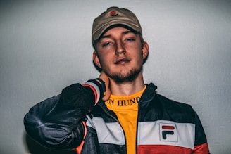 A person wearing a beige cap, a yellow shirt, and a black and white jacket with red details. The individual appears relaxed with one hand on the back of their neck and a calm expression on their face.