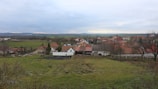 A small rural village landscape with scattered houses characterized by red-tiled roofs. The foreground includes grassy fields with a few bare trees, leading to a collection of houses surrounded by open fields. In the background, rolling hills and a cloudy sky complete the setting.