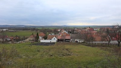 A small rural village landscape with scattered houses characterized by red-tiled roofs. The foreground includes grassy fields with a few bare trees, leading to a collection of houses surrounded by open fields. In the background, rolling hills and a cloudy sky complete the setting.