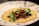 A corn tortilla topped with shredded meat, fresh cilantro leaves, and a red chili pepper. The tortilla is placed on a decorative plate with blue floral designs around the edge.