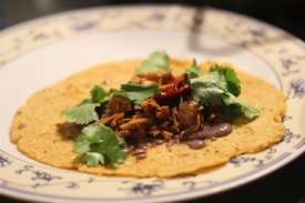 A corn tortilla topped with shredded meat, fresh cilantro leaves, and a red chili pepper. The tortilla is placed on a decorative plate with blue floral designs around the edge.