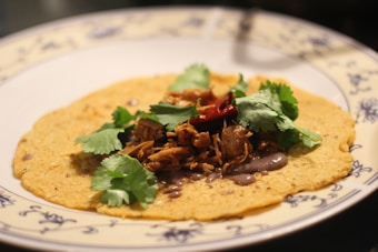 A corn tortilla topped with shredded meat, fresh cilantro leaves, and a red chili pepper. The tortilla is placed on a decorative plate with blue floral designs around the edge.