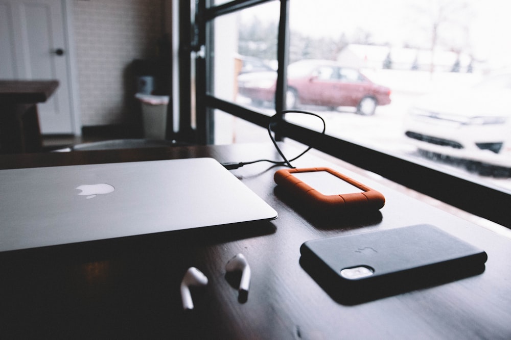 external hard drive plugged in MacBook near AirPods and iPhone X on table near window