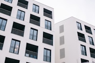 Two modern residential buildings featuring a minimalist design with white facades and dark window frames. Each building has multiple balconies with black railings, and the architecture emphasizes clean lines and geometric shapes.