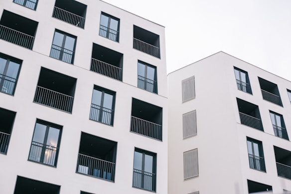 Two modern residential buildings featuring a minimalist design with white facades and dark window frames. Each building has multiple balconies with black railings, and the architecture emphasizes clean lines and geometric shapes.