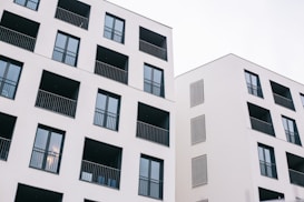 Two modern residential buildings featuring a minimalist design with white facades and dark window frames. Each building has multiple balconies with black railings, and the architecture emphasizes clean lines and geometric shapes.