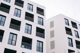 Two modern residential buildings featuring a minimalist design with white facades and dark window frames. Each building has multiple balconies with black railings, and the architecture emphasizes clean lines and geometric shapes.