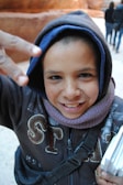 A young child is wearing a hooded jacket and scarf, smiling at the camera and making a peace sign with their fingers. The child has some marks on their face and is holding several papers or booklets. In the background, there are blurred outlines of rocky terrain and a couple of people walking away.