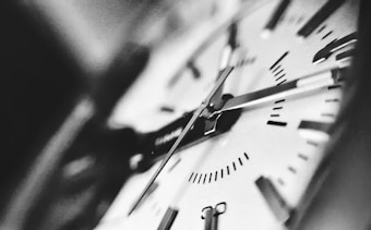 A close-up of an analog clock face with prominent hands and bold hour markers. The image is in black and white, highlighting the minute and hour hands and capturing the intricate details of the clock's design.