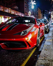 A sleek crimson luxury car parked on a city street at dusk, with soft urban lights reflecting off its polished surface.