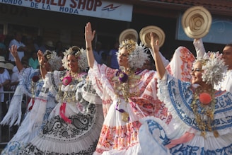 Three women are wearing traditional embroidered dresses adorned with floral and floral motifs. They have elaborate headpieces and are performing a dance with raised arms. The scene takes place outdoors with a background sign and a crowd observing the performance.