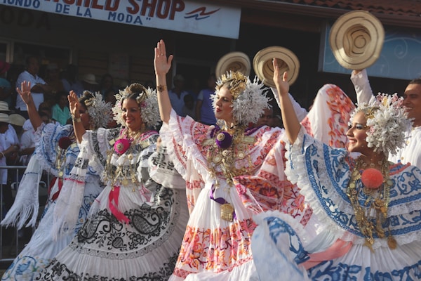 Three women are wearing traditional embroidered dresses adorned with floral and floral motifs. They have elaborate headpieces and are performing a dance with raised arms. The scene takes place outdoors with a background sign and a crowd observing the performance.